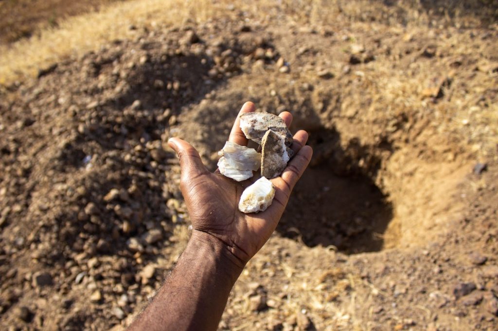 A hand holding rocks above a dirt-filled area with a shallow hole in the background.