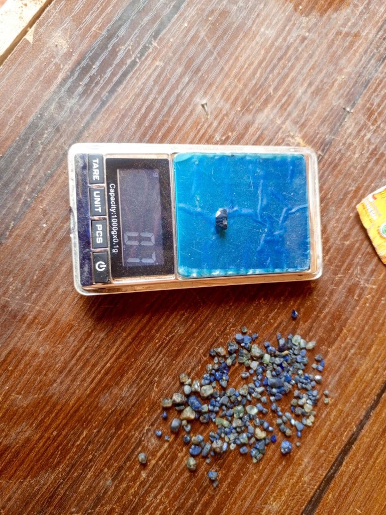 Digital scale weighing a small blue stone next to a pile of similar stones on a wooden surface.