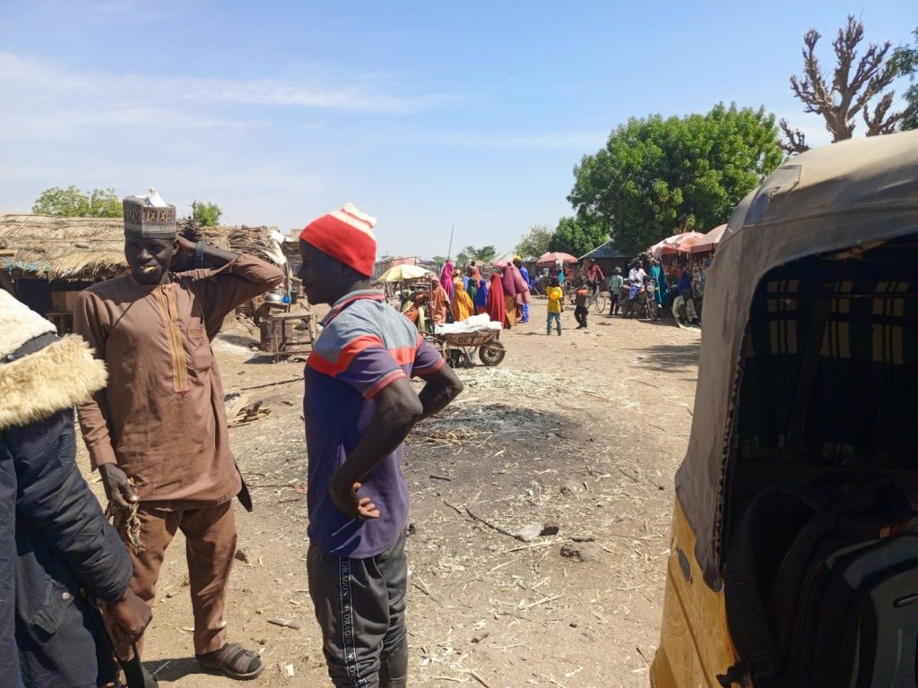 Outdoor market scene with people talking, stalls in the background, and a tree offering shade on a clear day.