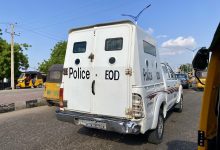 White police EOD vehicle on a busy street with tuk-tuks and cars, trees and a blue sky in the background.