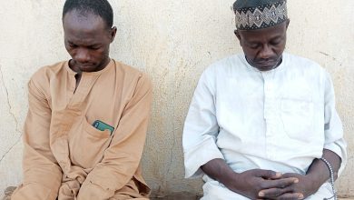 Two men, one in beige and the other in white with a cap, sit side by side against a textured wall, heads bowed, appearing contemplative.