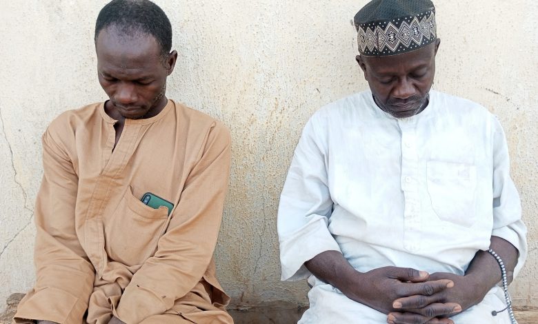 Two men, one in beige and the other in white with a cap, sit side by side against a textured wall, heads bowed, appearing contemplative.