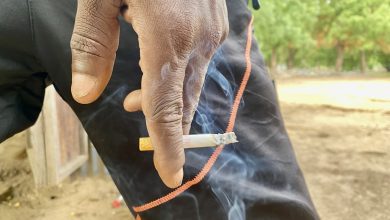 Hand holding a lit cigarette against black pants with orange trim, outdoors. Smoke wafts in the air.