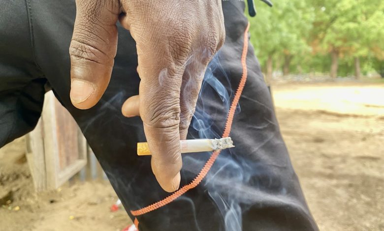 Hand holding a lit cigarette against black pants with orange trim, outdoors. Smoke wafts in the air.