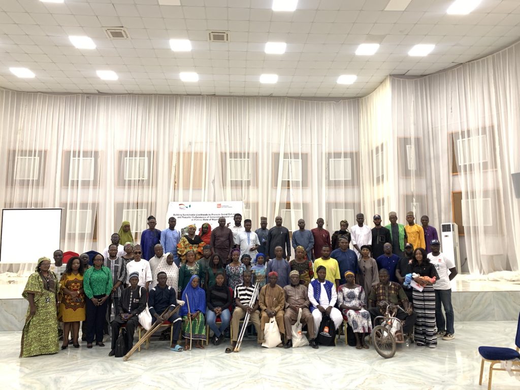 A large diverse group of people poses together in a bright room with a high ceiling, in front of a banner.