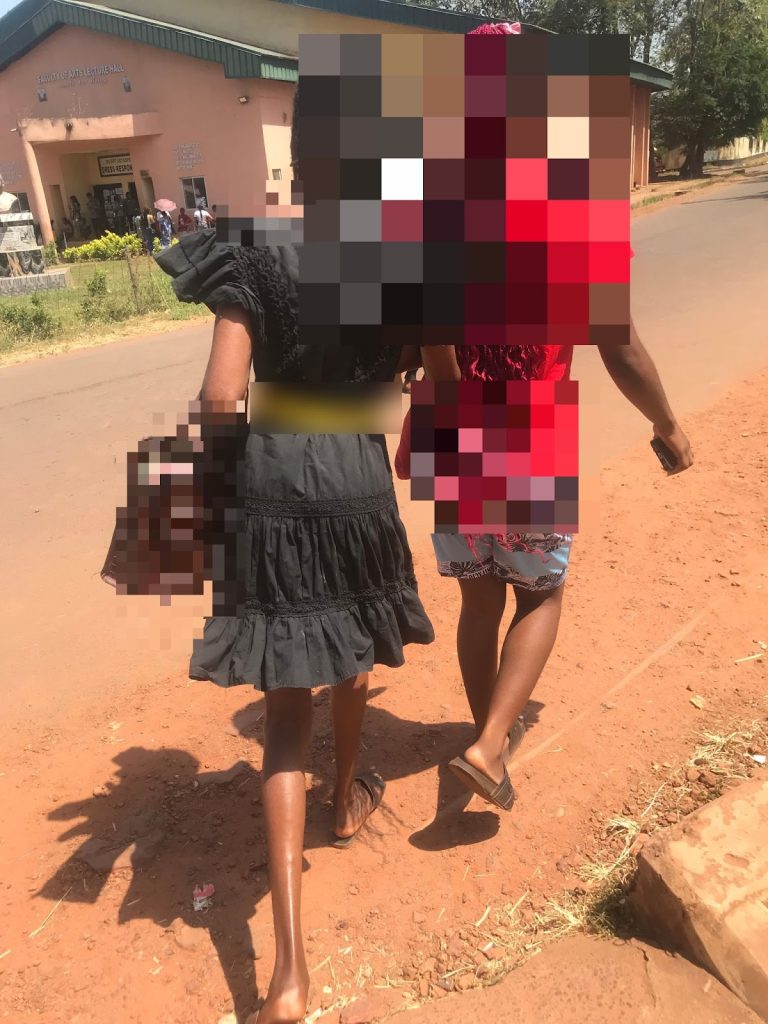 Two people walking on a dirt path towards a building, one in a black dress and the other in a red top.