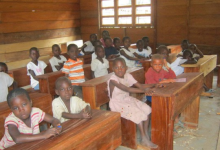 Children sitting at wooden desks in a classroom with wooden walls and windows, attentively looking ahead.