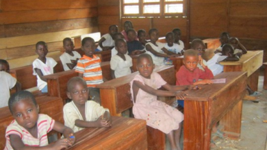 Children sitting at wooden desks in a classroom with wooden walls and windows, attentively looking ahead.