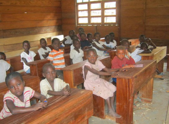 Children sitting at wooden desks in a classroom with wooden walls and windows, attentively looking ahead.