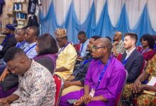 Audience attentively listening at a conference, people wearing lanyards and various cultural outfits, seated in red chairs.