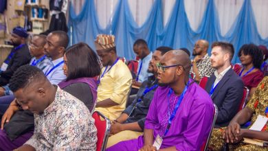 Audience attentively listening at a conference, people wearing lanyards and various cultural outfits, seated in red chairs.