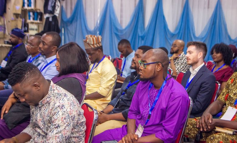 Audience attentively listening at a conference, people wearing lanyards and various cultural outfits, seated in red chairs.