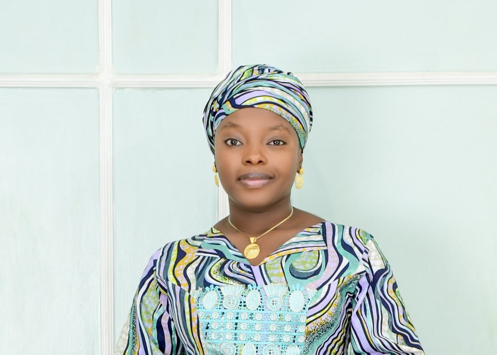 Woman in colorful patterned attire and headscarf, with a calm expression, standing in front of a light-colored wall.