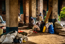 People sitting and standing with bags and bundles in a brick building's corridor, in warm sunlight.