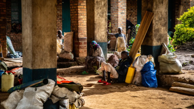 People sitting and standing with bags and bundles in a brick building's corridor, in warm sunlight.