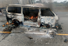 Burned-out van with flames inside, on a highway. Smoke rises, and a truck is visible in the background.