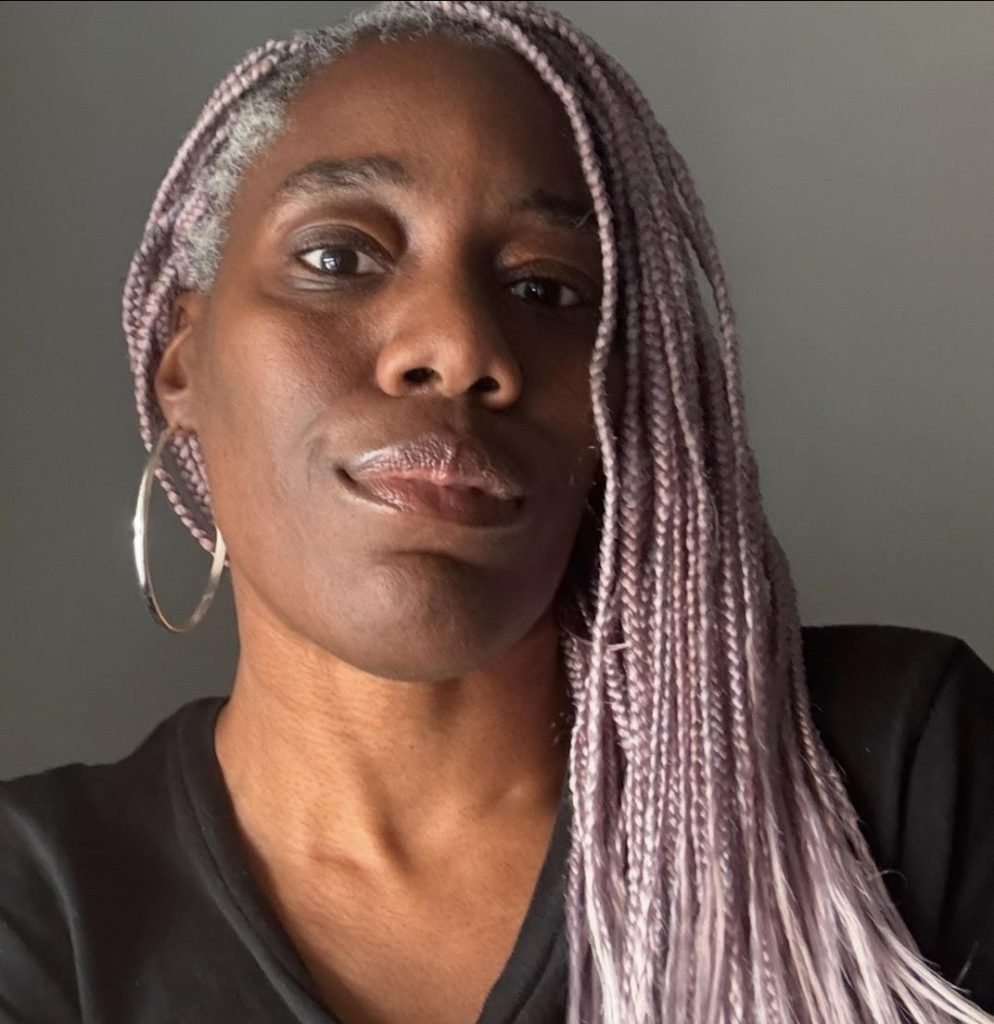 Person with long lavender braids and hoop earrings, wearing a black shirt, against a neutral background.