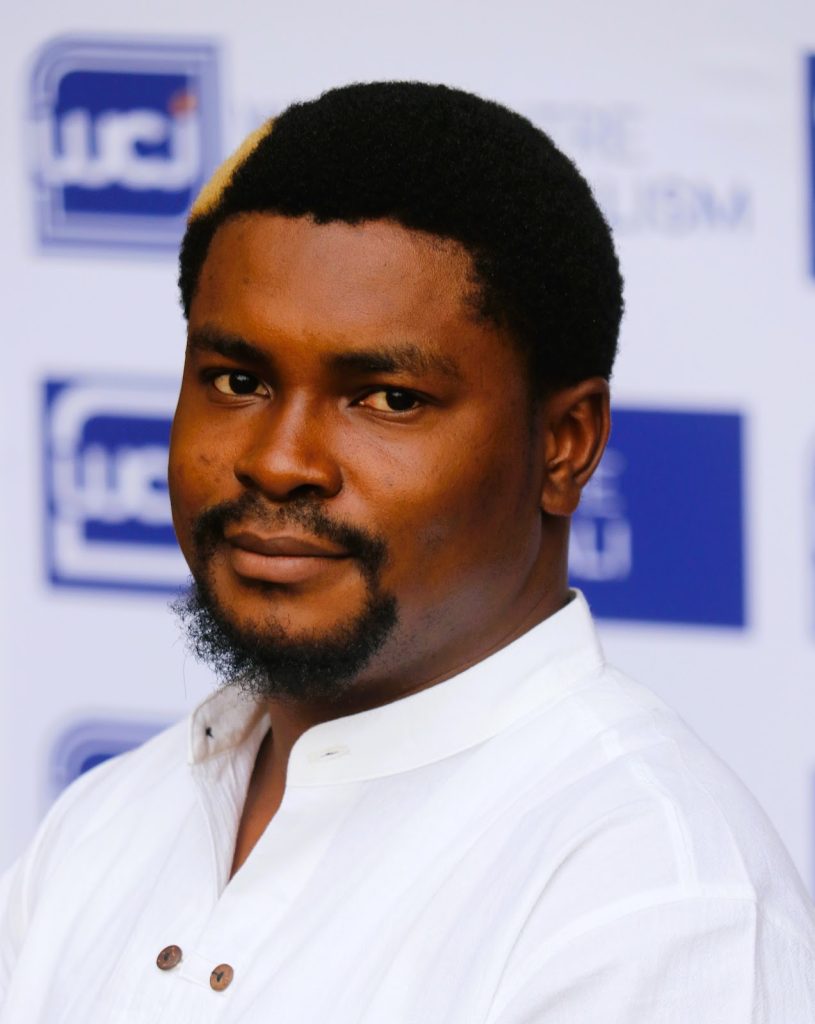 A person in a white shirt with a beard, standing against a background featuring repeated logo designs.