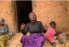 Three people sit outside a mud brick house. A woman gestures, another sits silently, and a child writes on paper.