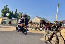 Three people ride a motorcycle on a sunny street, passing by a simple building with people standing outside.
