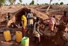 Children working in a rural area with tools and water containers, surrounded by dirt and trees.