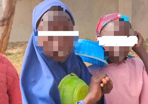 Two children standing, one in a blue hijab holding a green container, the other in a pink dress with a colorful cap.