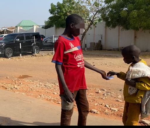 Two children exchanging a piece of paper on a sunny street, with parked cars and trees in the background.