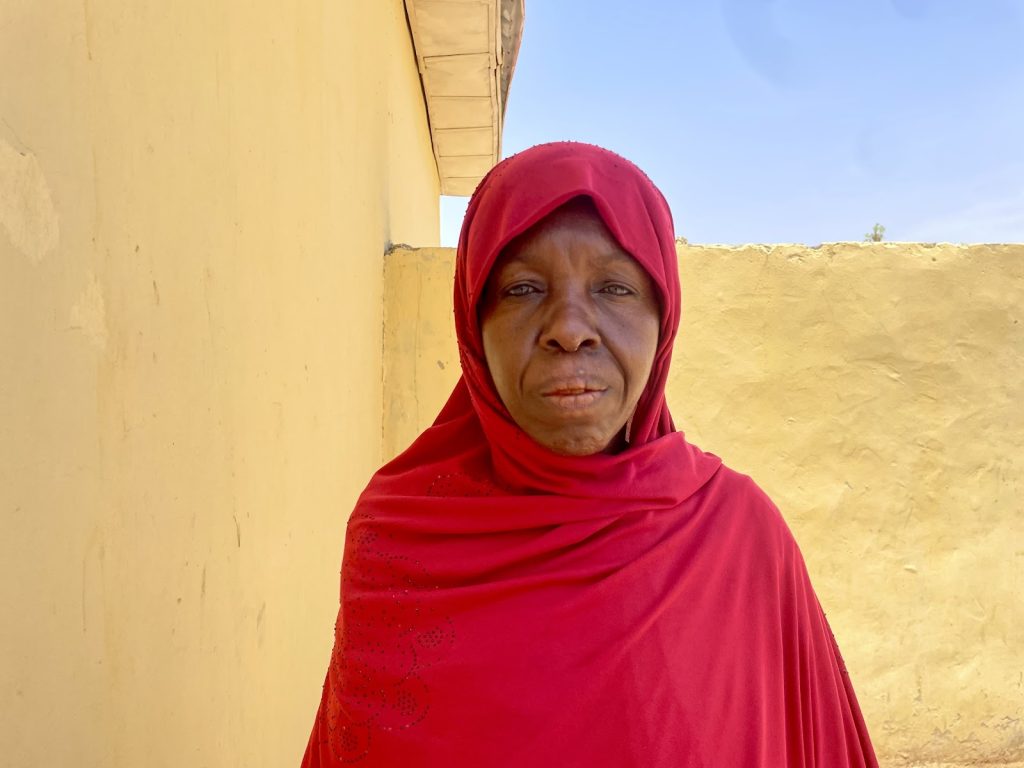 A person in a red hijab stands outside against a yellow wall, under a clear blue sky.