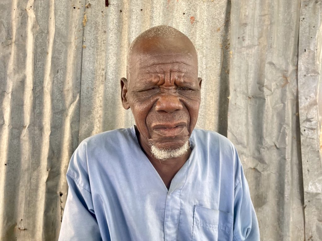 Elderly man in a light blue shirt stands against a corrugated metal wall, eyes closed, with a thoughtful expression.
