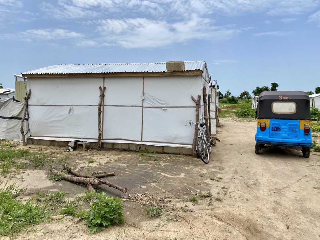 White tent structure next to a blue auto rickshaw and a bicycle on a dirt road under a cloudy sky.
