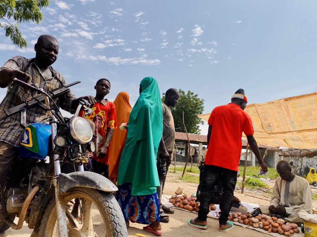 A group of people are gathered at an outdoor market, with onions displayed on the ground. A man on a motorcycle is in the foreground.