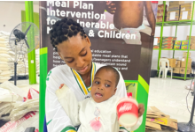 Woman holding a child in front of a "Meal Plan Intervention" poster inside a facility with shelves and bags.