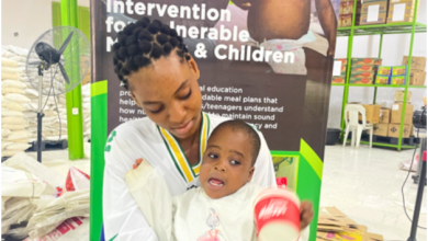 Woman holding a child in front of a "Meal Plan Intervention" poster inside a facility with shelves and bags.