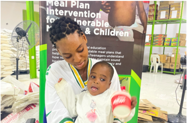 Woman holding a child in front of a "Meal Plan Intervention" poster inside a facility with shelves and bags.