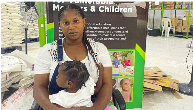 A woman sits with a child in her lap in front of a sign about resources for vulnerable mothers and children, surrounded by bags and boxes.