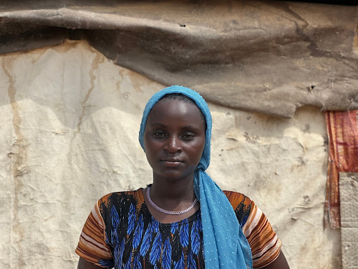 A person wearing a headscarf and colorful clothing stands in front of a textured, rustic wall.
