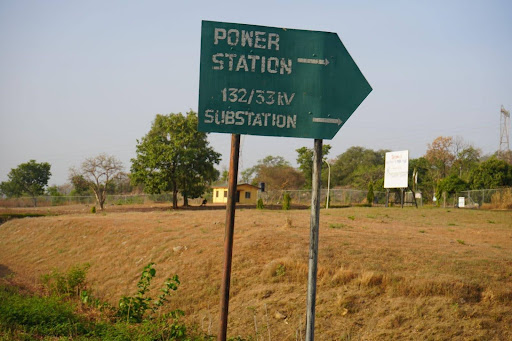 Sign pointing to power station and substation, 132/33kV, in a grassy field with scattered trees and a distant building.