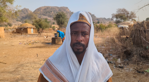 Man in traditional clothing stands outside, with huts and hills in the background.