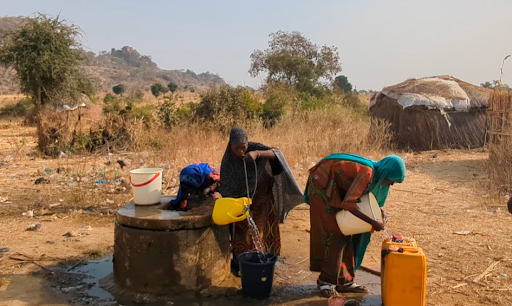 Two women collect water from a well in a rural area, with dry grass, a hut, and hills in the background.