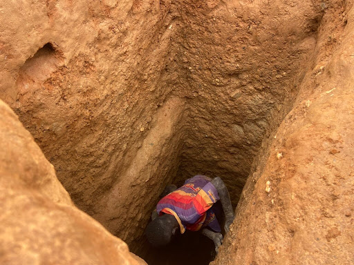 Person in a brightly colored shirt digging in a deep, narrow dirt trench.