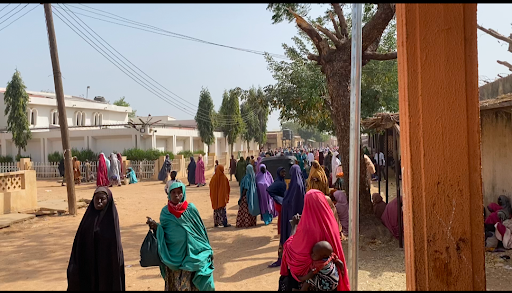 People in colorful clothing gather on a street lined with trees and houses, under clear skies.