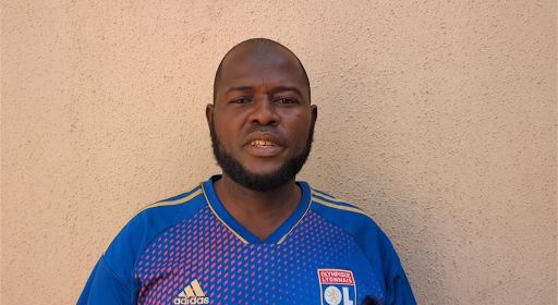 A man with a beard stands against a textured wall, wearing a blue sports jersey with red and yellow accents.