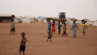 Children and women carrying wood walk in a dirt area near simple structures and tents in an open field.