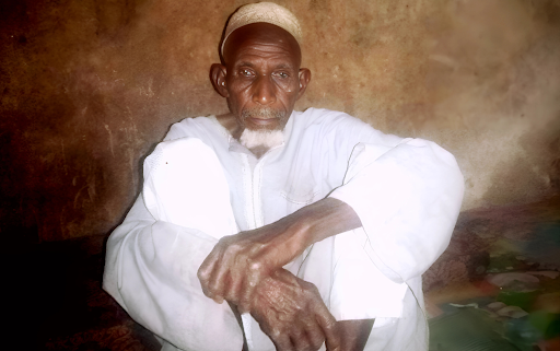 Elderly man in white clothes and a cap, seated against a textured wall, with hands resting on his knee.