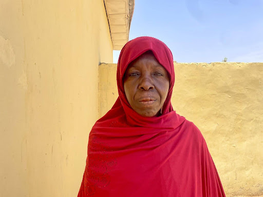 Person wearing a red headscarf stands against a beige wall with blue sky in the background.