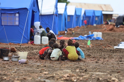 Children sit in a circle on muddy ground near blue tents in a refugee camp.
