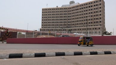 A yellow auto rickshaw drives past a large, multi-story building and a red wall on a clear day.