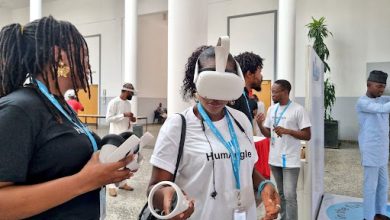 Person wearing a VR headset at a tech event, surrounded by others holding VR equipment, in a modern indoor space.