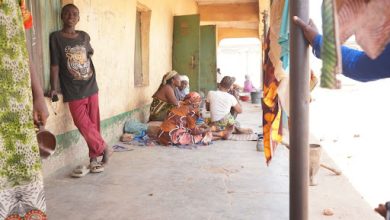 People gathered on a porch in casual attire, with colorful fabrics hanging, engaging in conversation on a sunny day.