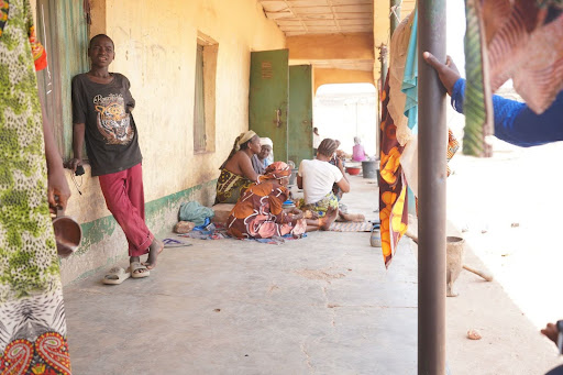 People gathered on a porch in casual attire, with colorful fabrics hanging, engaging in conversation on a sunny day.